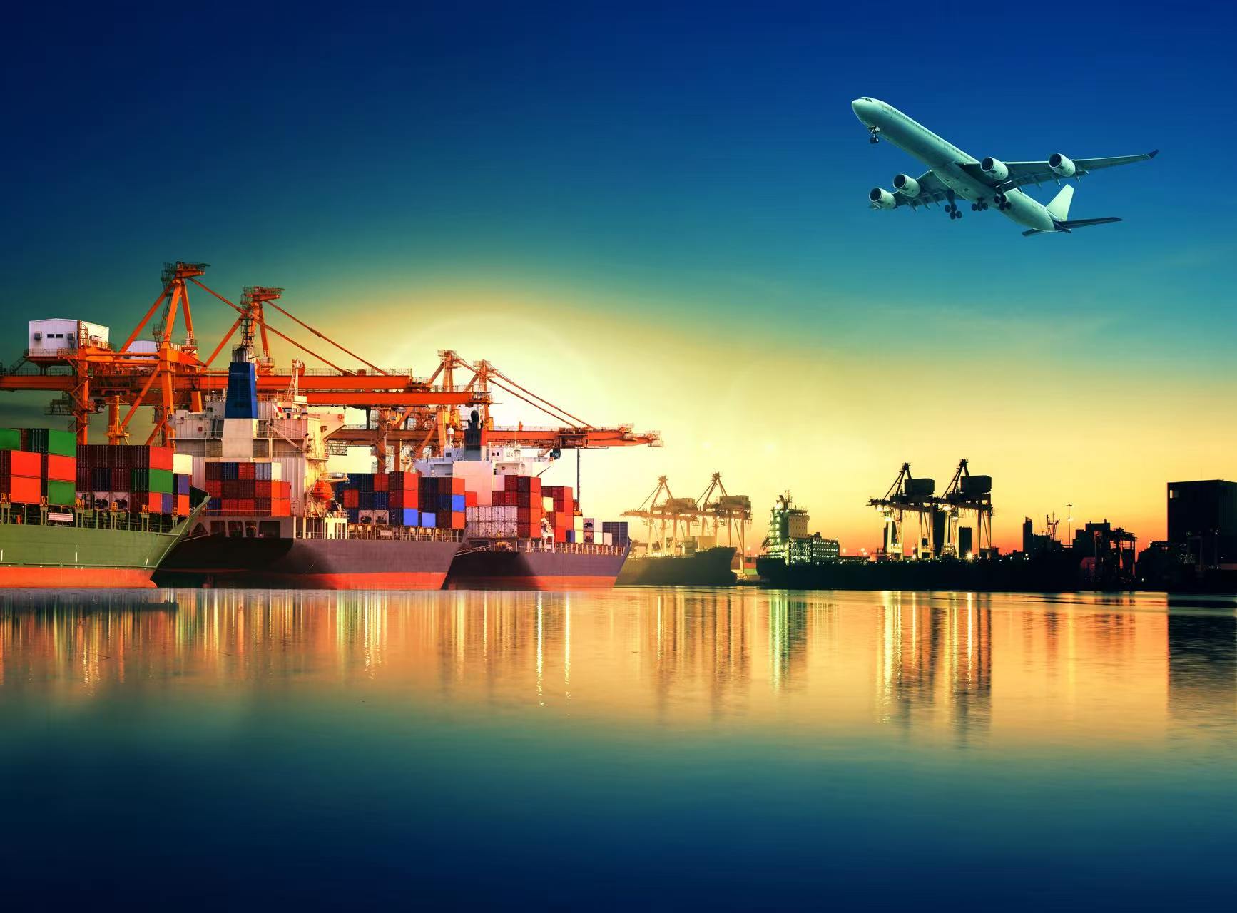 Cargo vessels and container cranes at a port at sunset, with an aircraft overhead